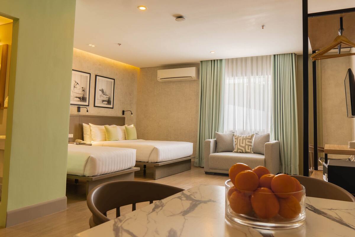 A hotel room with two beds, a sofa, a dining table with a bowl of fruit, and a TV.