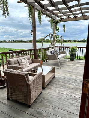 Una terraza de madera con muebles de mimbre, una hamaca y vista al lago.