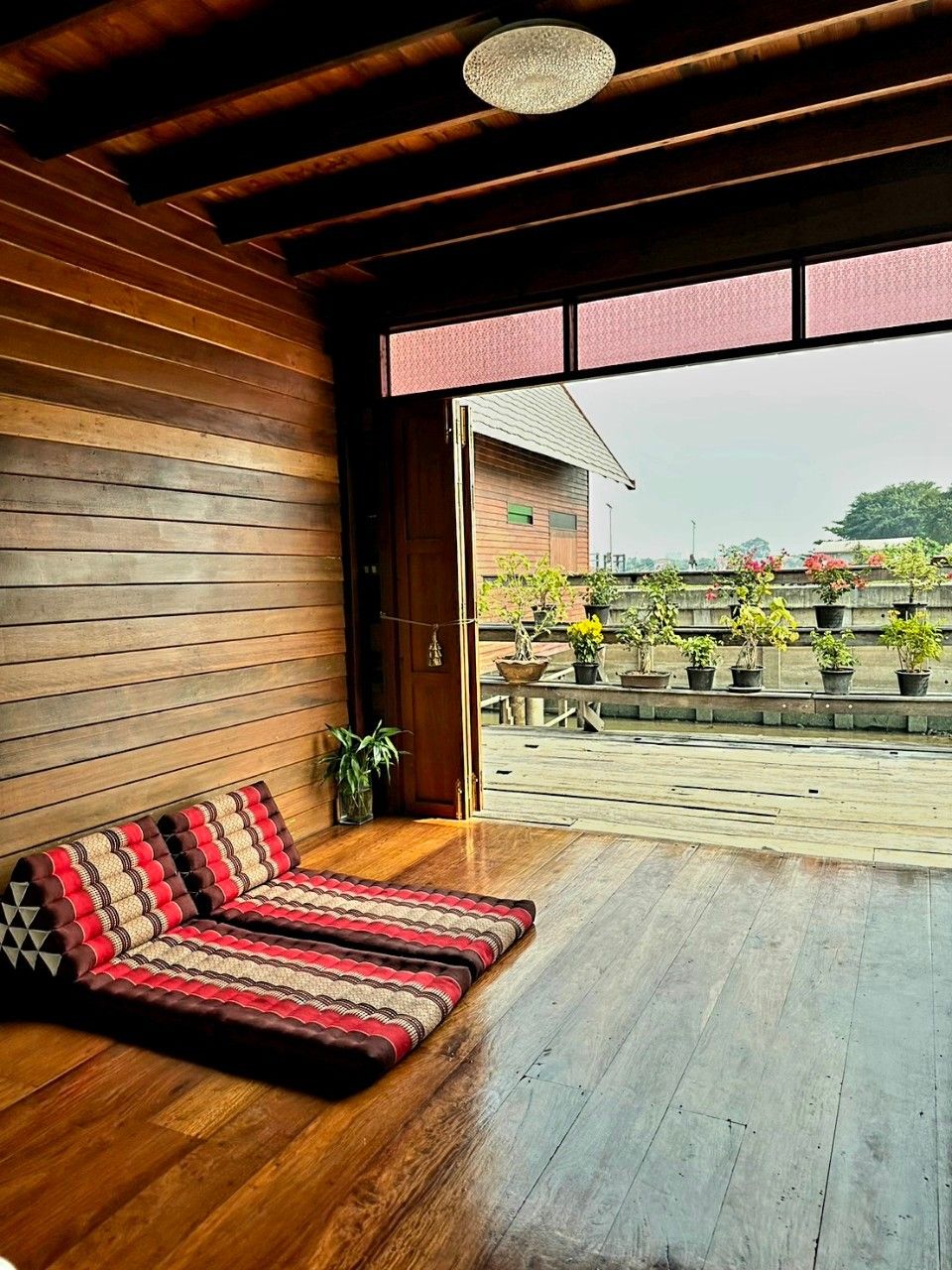 A room with a wooden floor, a bench with a patterned cushion, and a view of a rooftop garden with potted plants.