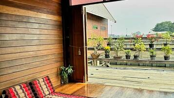A room with a wooden floor, a bench with a patterned cushion, and a view of a rooftop garden with potted plants.