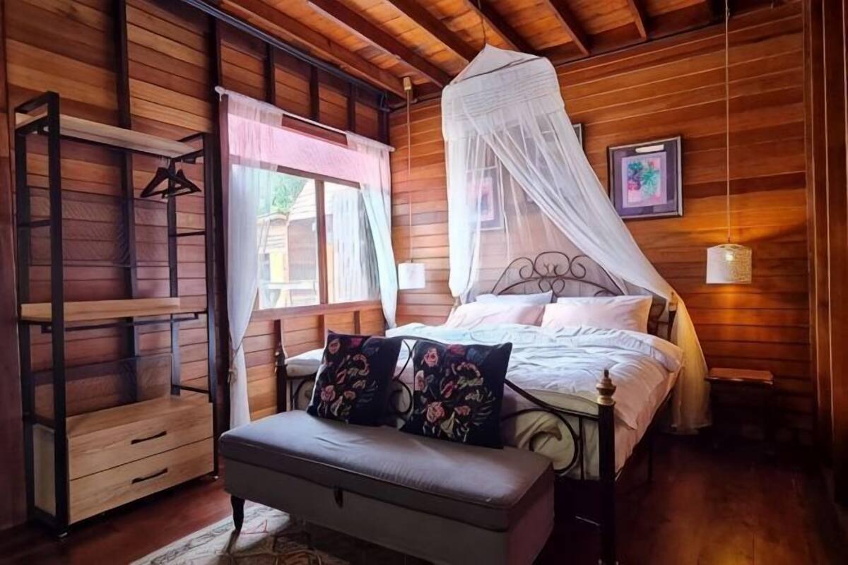 Une pièce en bois avec un lit, un banc, une chaise suspendue et une fenêtre avec des rideaux.