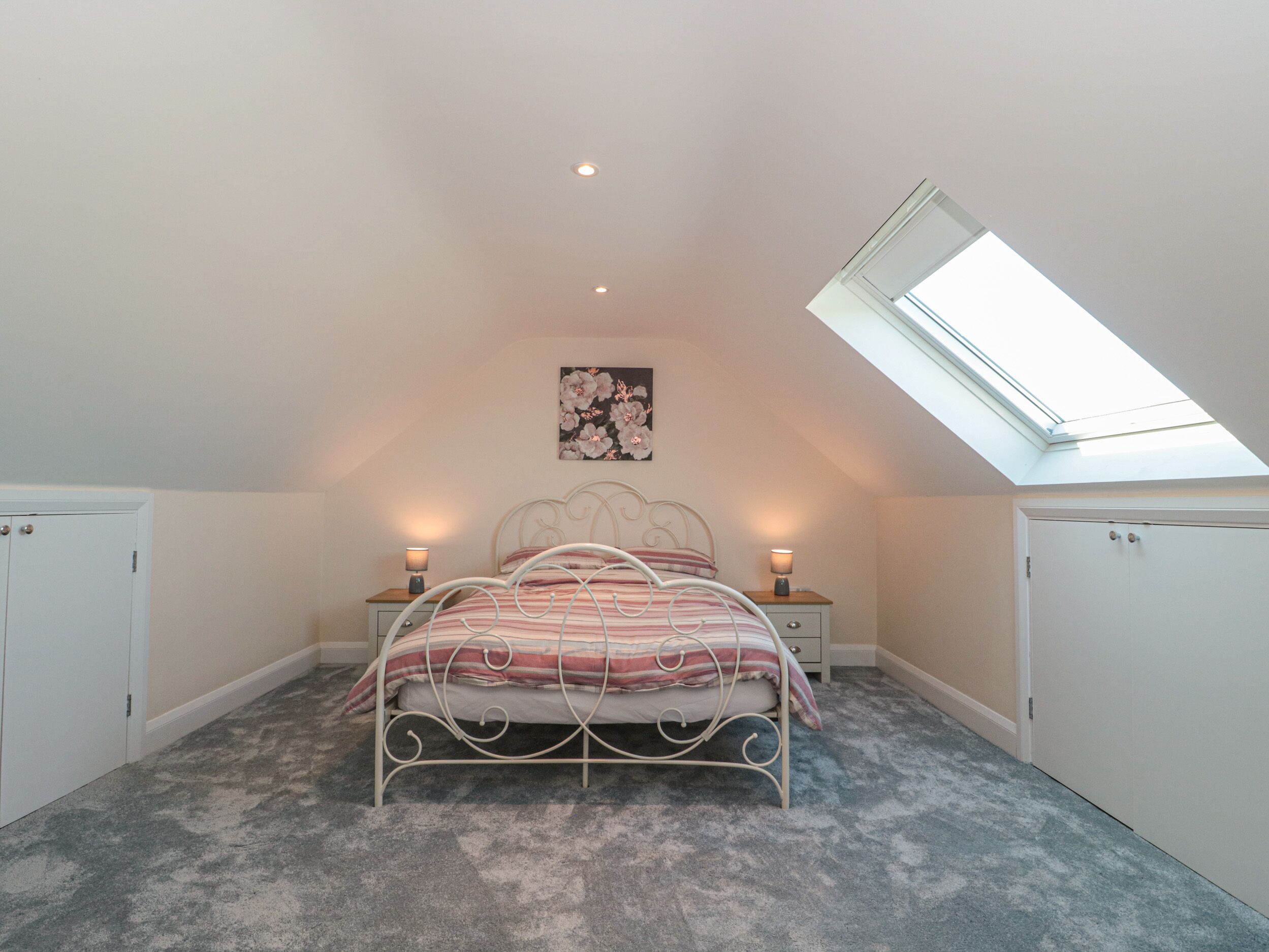 A bedroom with a skylight, a bed with a striped blanket, two bedside tables with lamps, and a floral artwork on the wall.