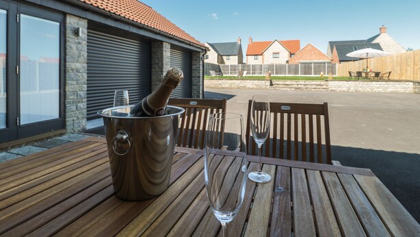 A wooden table with a champagne bottle and two glasses, set outdoors with a view of houses and a clear sky. - Homeland (Somerton)