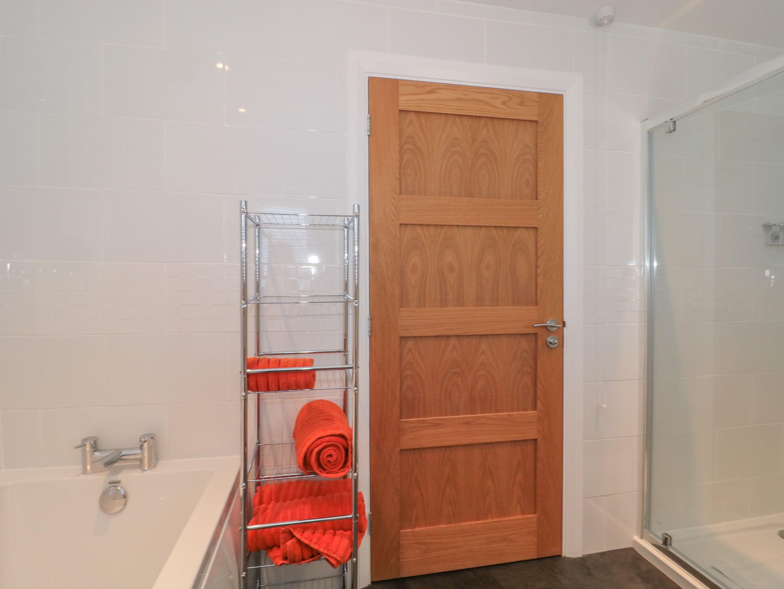 Un baño con puerta de madera, cabina de ducha de vidrio y un perchero de metal con toallas rojas.