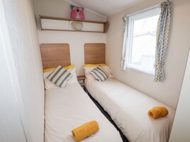 Une petite chambre accueillante avec deux lits simples, une fenêtre ornée de rideaux à motifs et une étagère avec des coussins décoratifs.