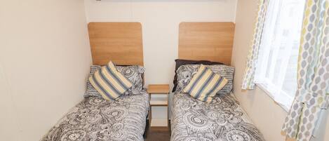 A small room with two single beds, a shelf with books, and a window with patterned curtains.