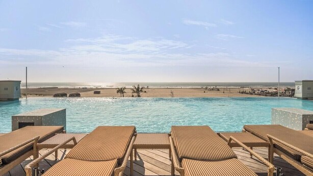 A pool with lounge chairs, a beach in the background, and a clear sky. - Residence 101 at The Grand (Wildwood)