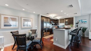 A modern kitchen with dark cabinetry, a central island, and a dining area with wicker chairs. - Residence 101 at The Grand (Wildwood)