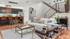 Una sala de estar moderna con sofá, mesa de centro, sillas, una escalera y un área de cocina con taburetes de bar.