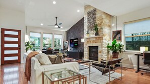 A spacious living room with a stone fireplace, a flat-screen TV, and a large window offering a view of the outdoors.