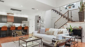 A modern living room with a sofa, coffee table, and chairs, a staircase, and a kitchen area with bar stools.