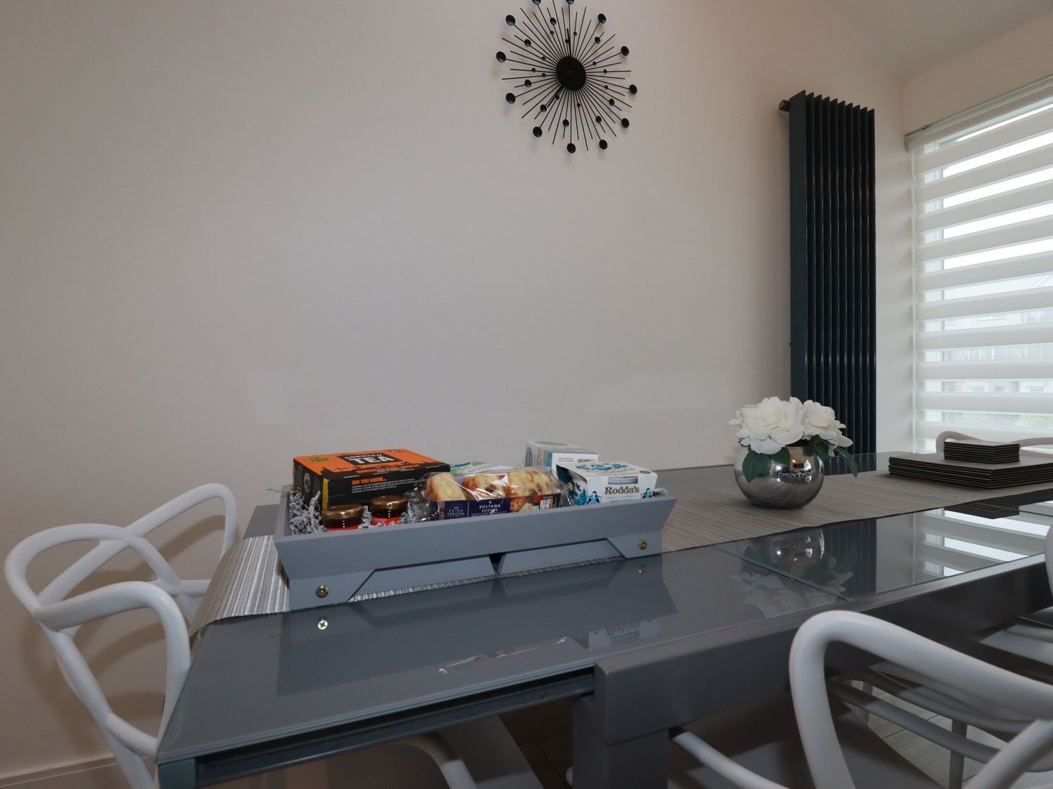 A modern dining area with a glass table, white chairs, a decorative wall piece, and a window with blinds.
