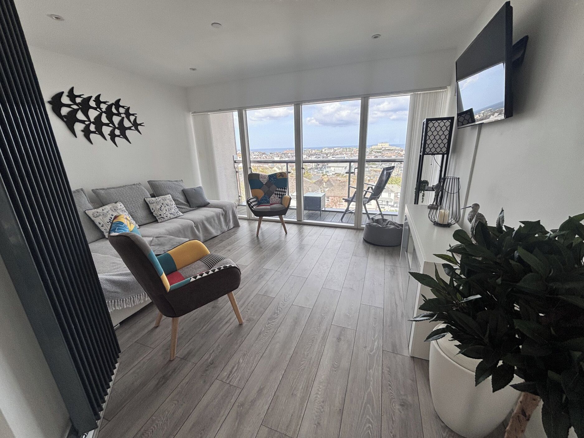 A modern living room with a grey sofa, a colorful armchair, a large window with a city view, and a potted plant.