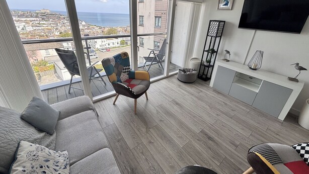 A modern living room with a large window, a sofa, a chair, a TV, and a small plant on a shelf. - 10 Quay Court (Newquay)