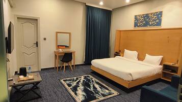 A hotel room with a bed, a desk, a chair, a small table, a mirror, a blue curtain, and a painting on the wall.