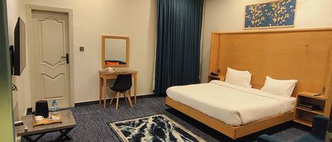 A hotel room with a bed, a desk, a chair, a small table, a mirror, a blue curtain, and a painting on the wall.