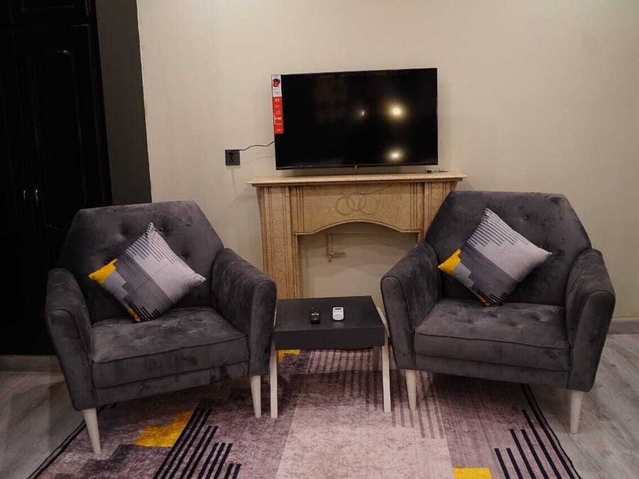 Een moderne woonkamer met twee grijze fauteuils, een klein zwart bijzettafeltje en een flatscreen televisie bevestigd aan een houten kast.