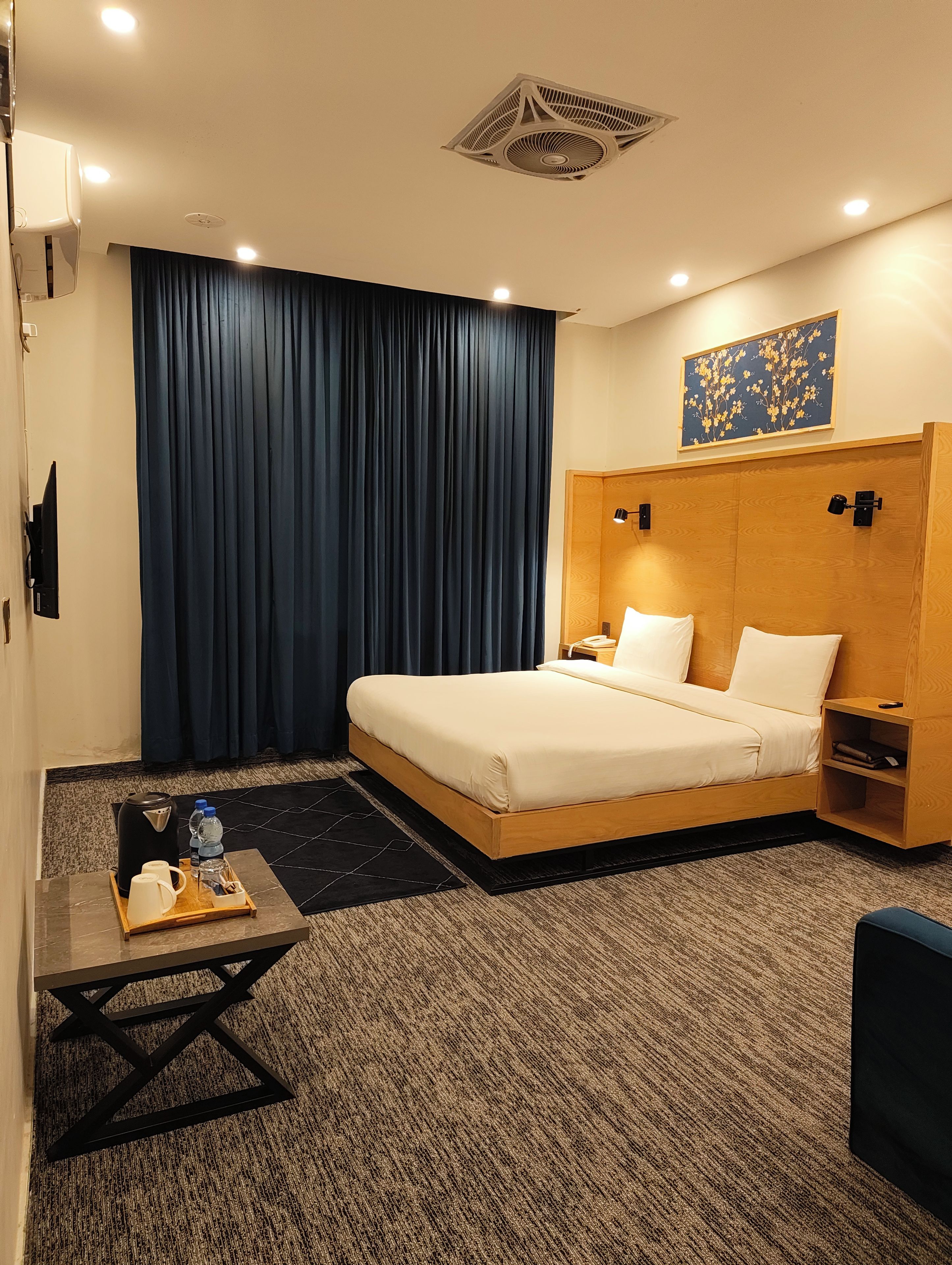 Une chambre d’hôtel comprenant un lit, une table de chevet, une machine à café, un plateau avec une bouilloire et des bouteilles d’eau, des rideaux occultants, une œuvre d’art accrochée au mur et un sol recouvert de moquette.