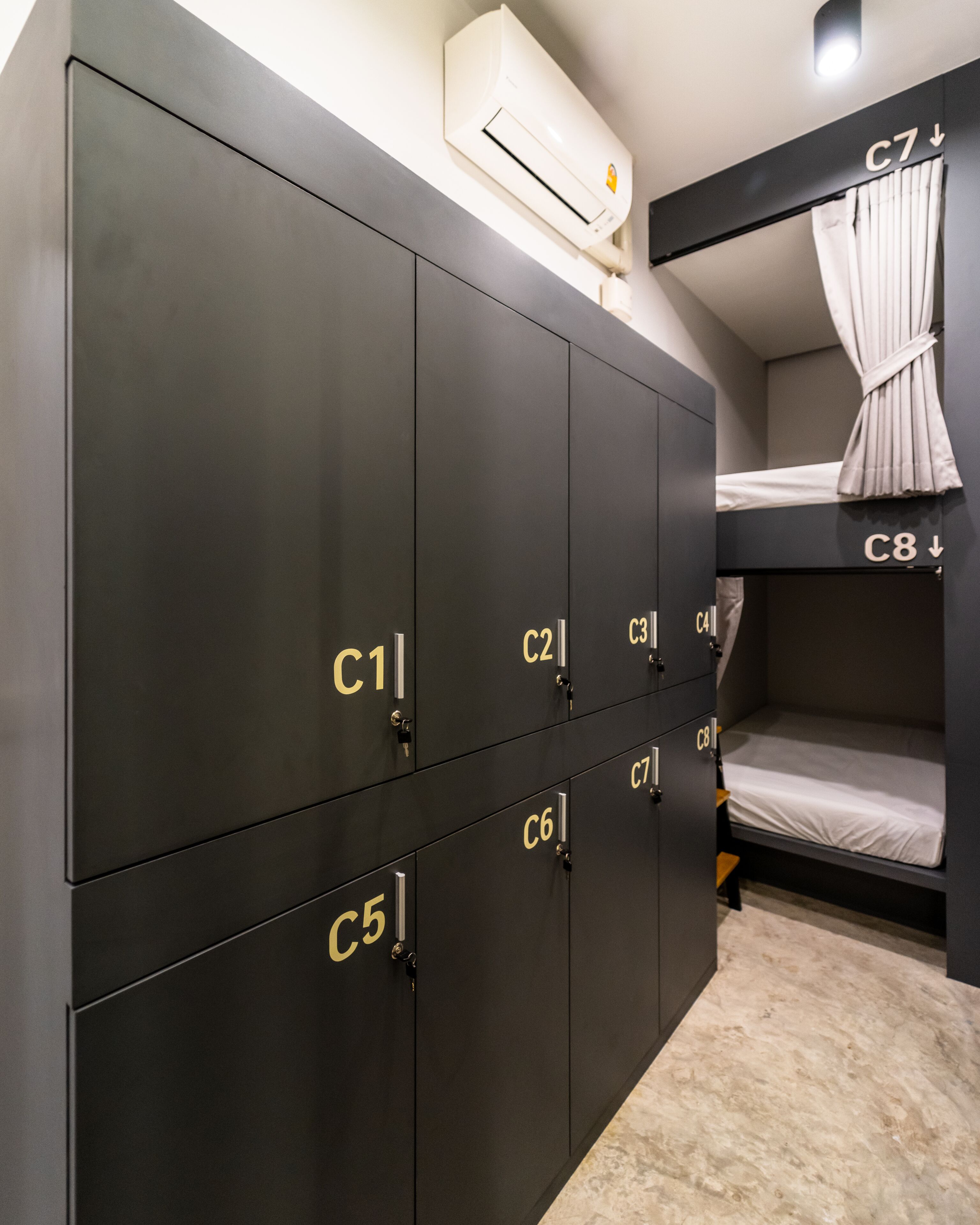 Une chambre de dortoir avec des lits superposés portant les numéros C7 et C8, une armoire avec plusieurs compartiments et un appareil de climatisation fixé au mur.