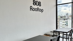 A modern interior with a black table and chairs, a white wall with the number 808 Rooftop, and a large window offering a city view. - PIER 808 Hostel (Bangkok)