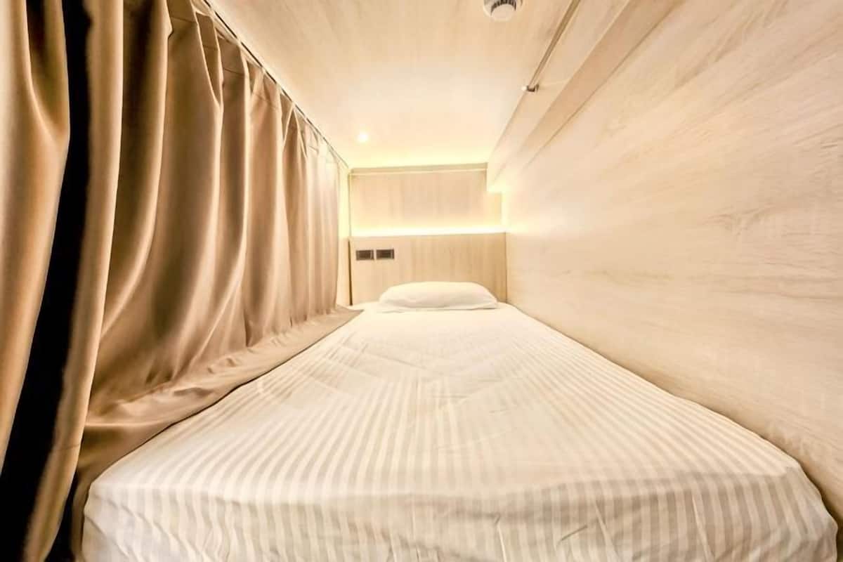 A narrow hotel room with a single bed, a headboard with built-in lighting, and a curtain on one side.