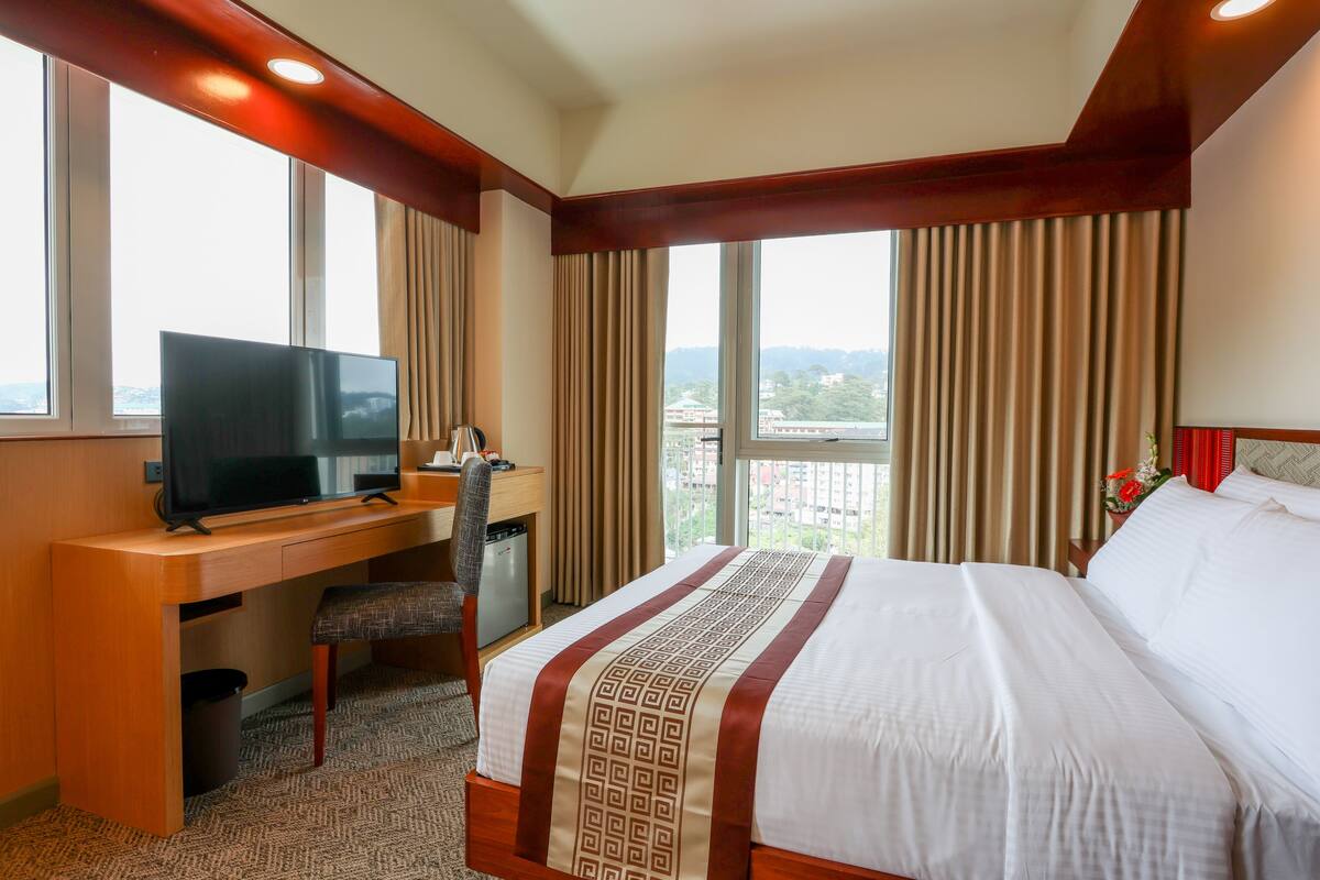 Una habitación de hotel con una cama, un escritorio con televisor y vistas a la ciudad a través de amplias ventanas.