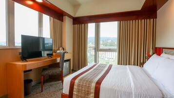 A hotel room with a bed, a desk with a television, and a view of the city through large windows.