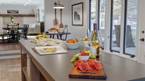 A modern kitchen with a long counter, a dining area in the background, and a view of the outdoors through large windows.