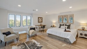 A spacious bedroom with a large bed, a desk, and a chair. There are multiple windows providing natural light and a view of the outdoors.