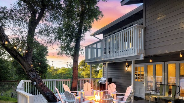 A deck with Adirondack chairs, a fire pit, and a grill, overlooking a house with a balcony.