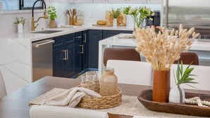 A modern kitchen with dark blue cabinets, white countertops, and stainless steel appliances. There are various kitchen utensils, a bread loaf, and decorative plants on the counter. - Element by AvantStay | Pool, Firepit, Game Room (Scottsdale)