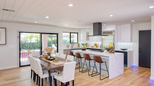 A modern kitchen with white cabinetry, a central island with bar stools, and a dining area with a wooden table and white chairs. - Element by AvantStay | Pool, Firepit, Game Room (Scottsdale)