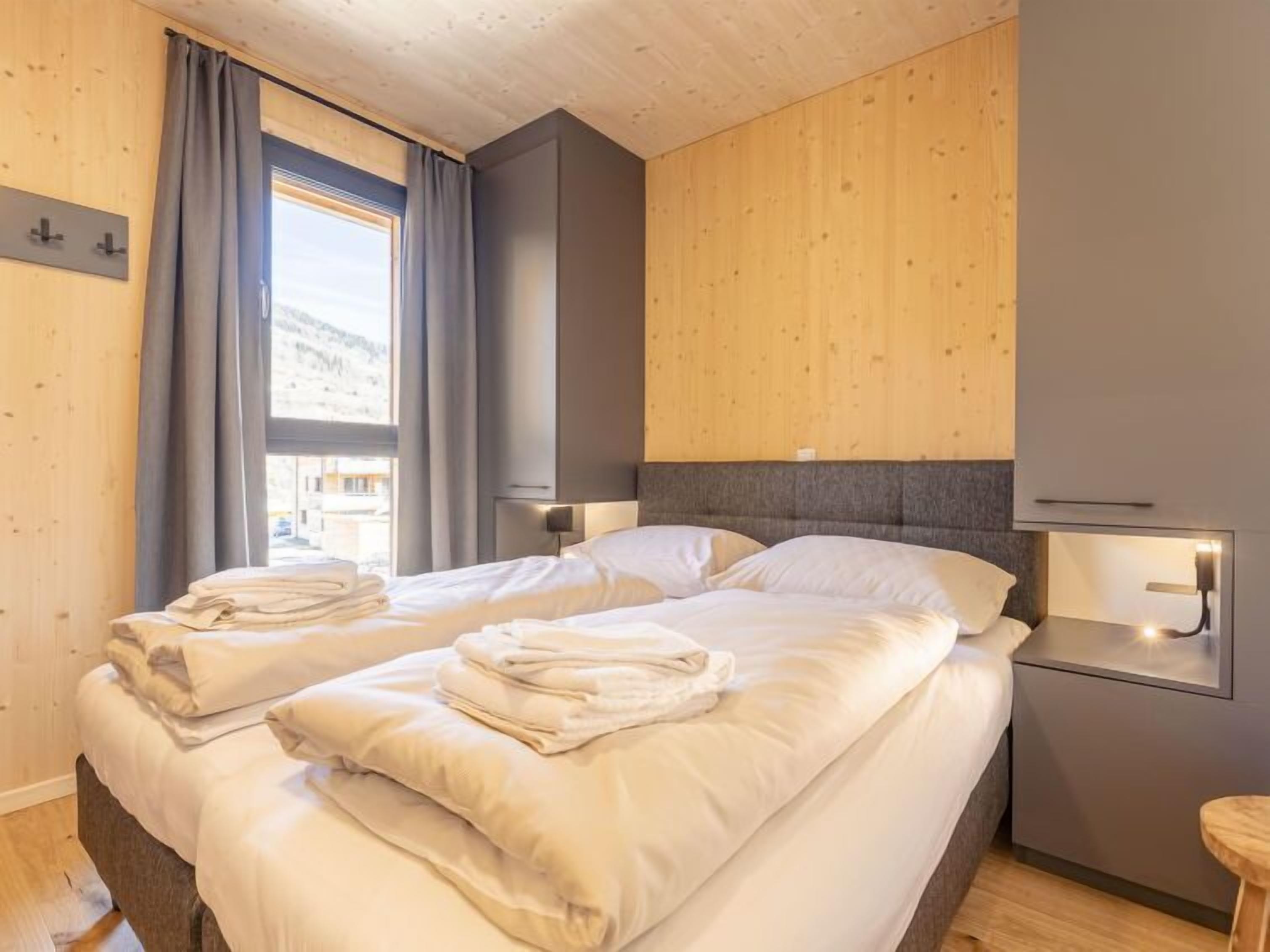 A modern hotel room with two beds, wooden paneling, a large window with curtains, and bedside lamps.