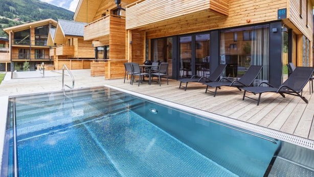 Una cabaña de madera con piscina, mobiliario exterior y montañas al fondo.