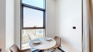 Une chambre d’angle avec une table à manger dressée pour deux personnes, une fenêtre donnant sur un paysage urbain et une porte.