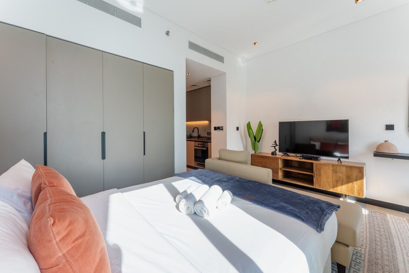 Una camera d'albergo moderna con un ampio letto, una TV, un mobiletto in legno e una piccola cucina sullo sfondo.