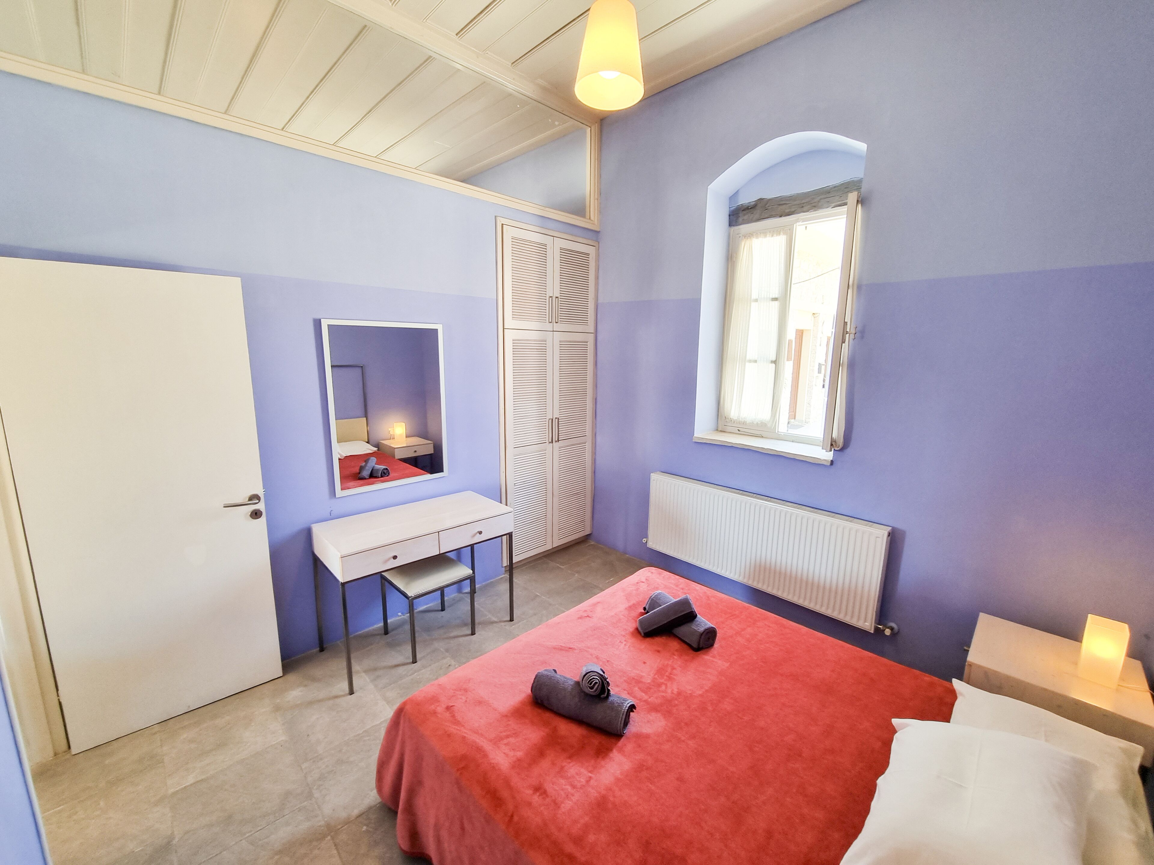 A bedroom with a bed, a desk, a chair, a wardrobe, a radiator, and a window.