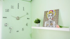 A clock with numbers 1-12 and a second hand, a painting of a person with a colorful headscarf, a small potted plant, and a pink flower on a white shelf. - Palm Beach House - Sea and Beach View (Montesilvano)