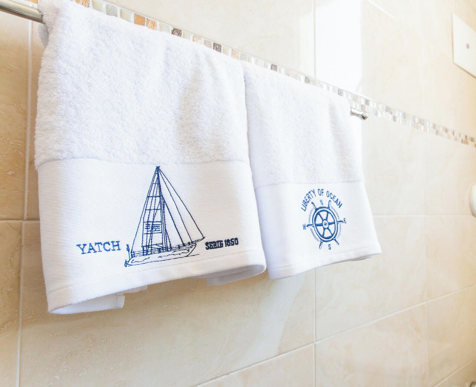 Two white towels hanging on a bathroom rack with nautical designs and text.
