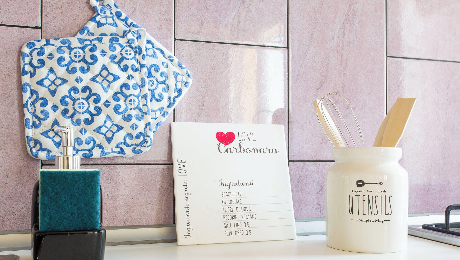 Un bancone cucina con un tovagliolo azzurro e bianco, una tazza bianca con posate e un menu che elenca gli ingredienti.