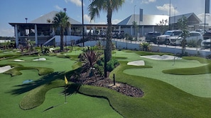 A golf course with a modern building in the background, palm trees, and a clear sky.