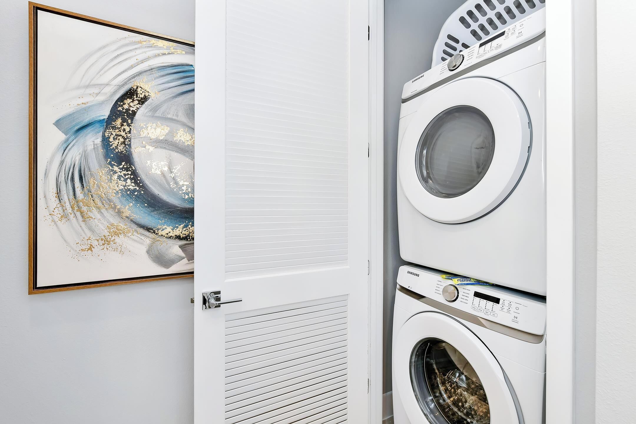 A laundry room with stacked washing machines and dryers, a door with a louvered vent, and a framed abstract painting on the wall.
