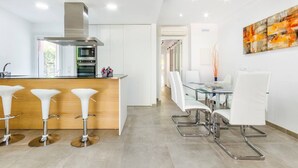 Una cocina moderna con una isla central, taburetes de bar blancos y un comedor con mesa de vidrio y sillas blancas.