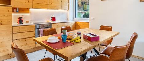 A modern kitchen with wooden cabinets, a dining table with chairs, and a view of a forest through a window.