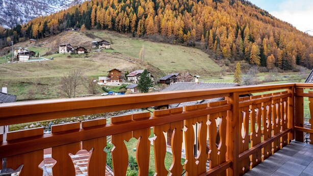 Une balustrade en bois surplombe un paysage montagneux, avec des maisons et un feuillage automnal.