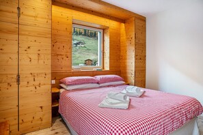 Une pièce de cabane en bois comprenant un lit, une literie à carreaux, une fenêtre donnant sur une maison et des arbres, ainsi qu’une armoire en bois.