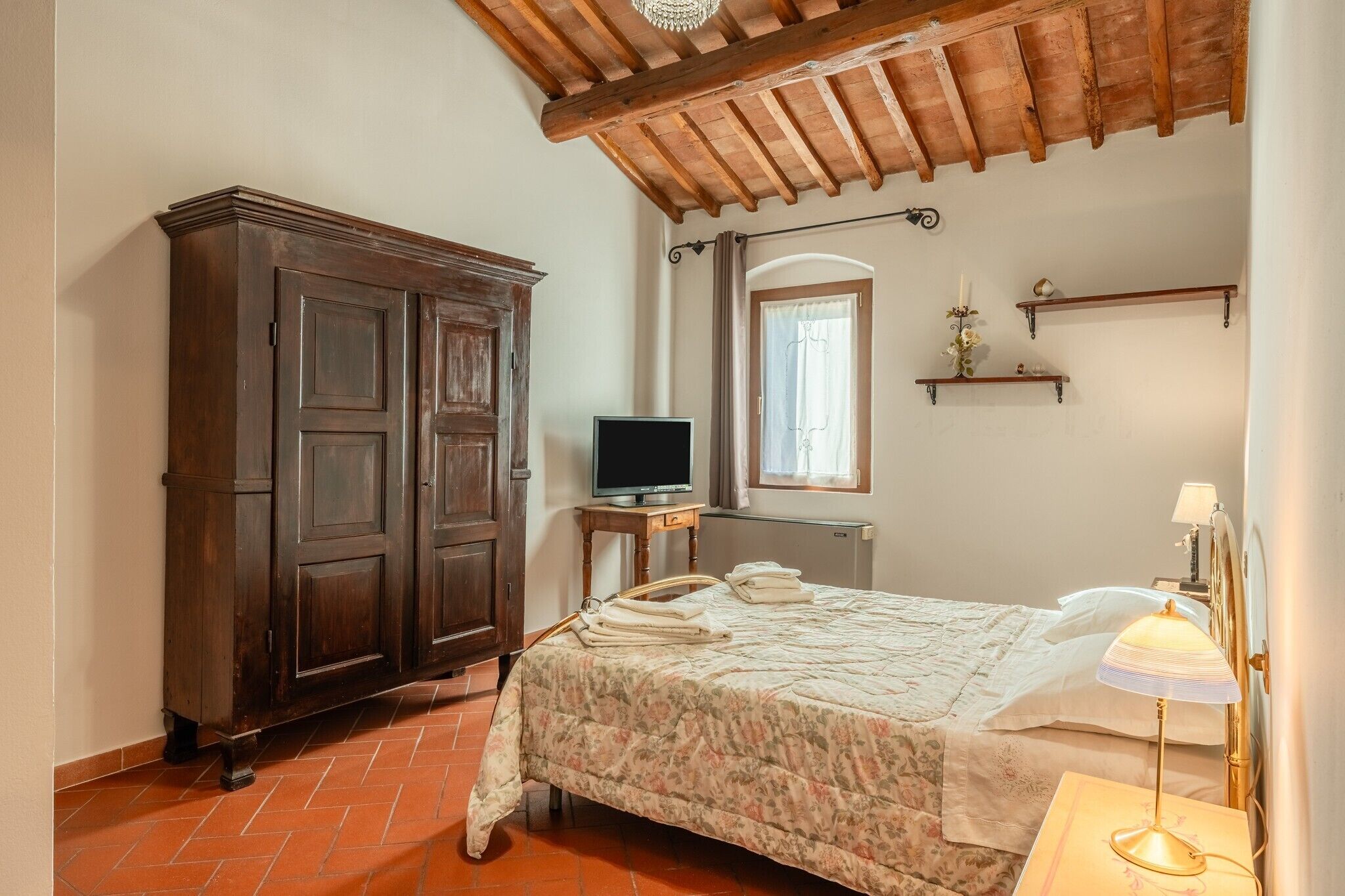 Une chambre avec une armoire en bois, un téléviseur posé sur une petite table, un lit recouvert d’une couvre-lit à motifs floraux, une table de chevet avec une lampe et une fenêtre avec des rideaux.