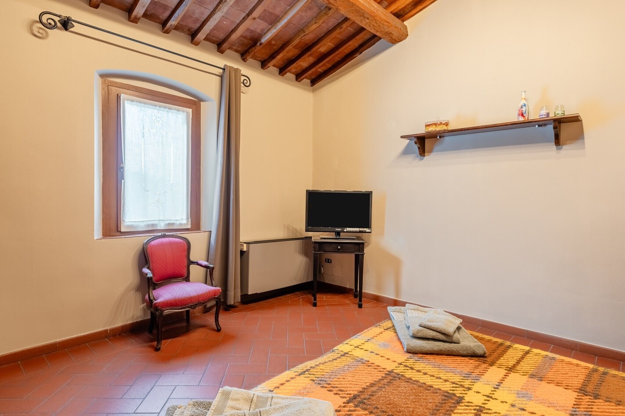 Une pièce comprenant une télévision, une chaise, une fenêtre avec des rideaux, un radiateur, une étagère avec des articles de toilette et un tapis à motifs.