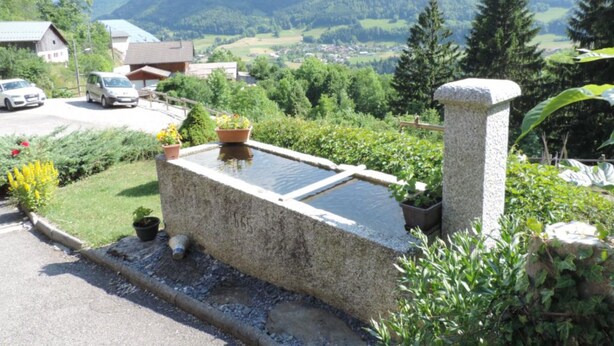 Une structure en pierre agrémentée d’un élément aquatique, entourée de verdure et dominant un paysage montagneux.