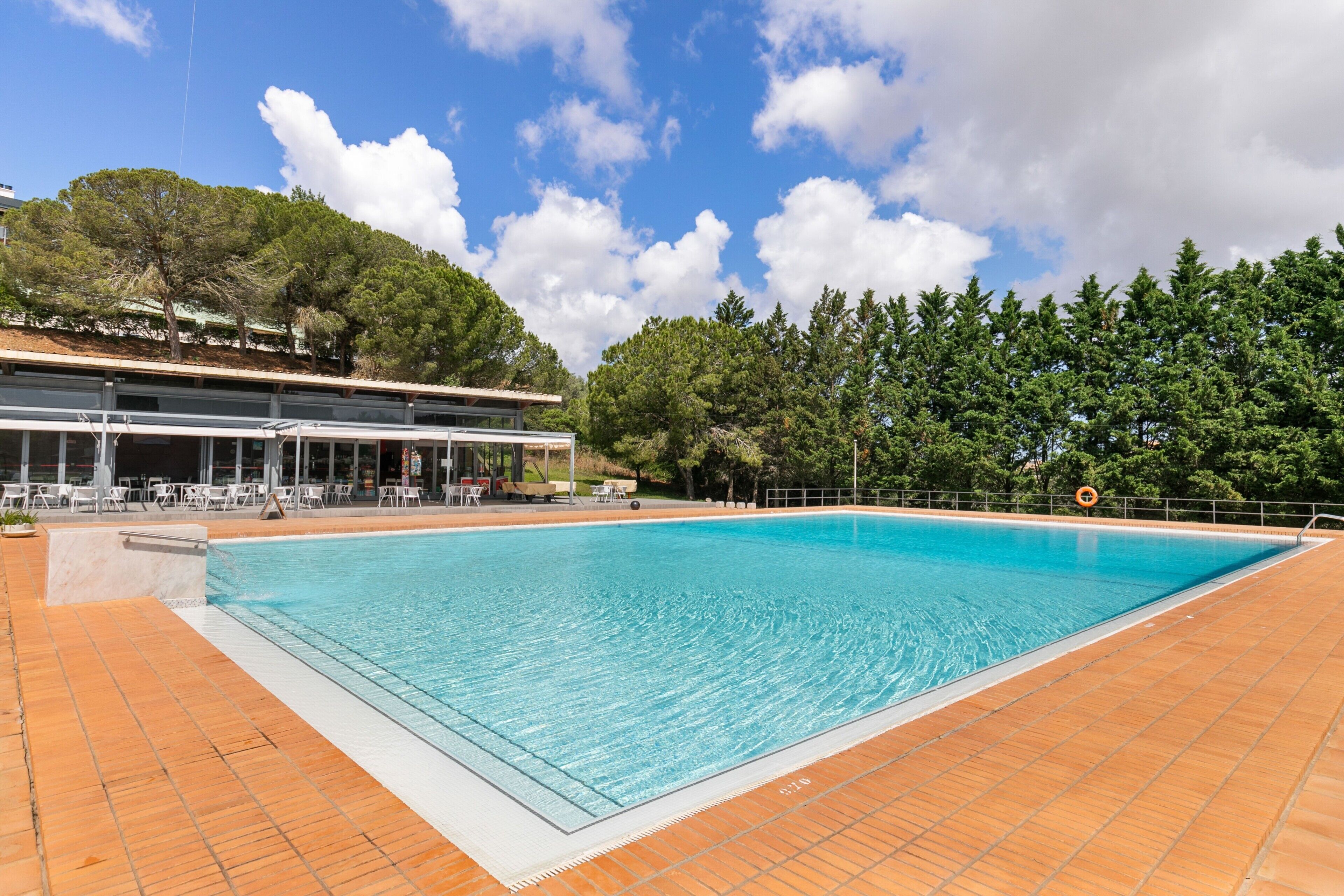 Una piscina con plataforma de baldosas, rodeada de árboles y un edificio con asientos al aire libre.